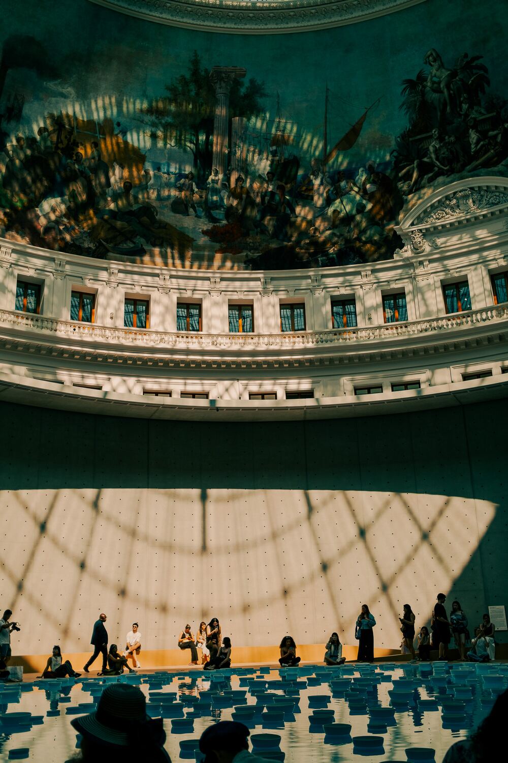 Visitors beneath the painted dome of the Bourse de Commerce in Paris, sunlight casting a lattice of shadow across the walls.