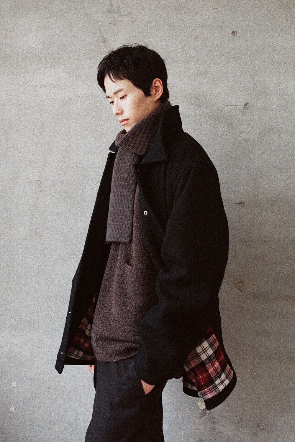 Horlisun 26 F/W lookbook: a man in a dark wool coat over a brown knit against a concrete wall.