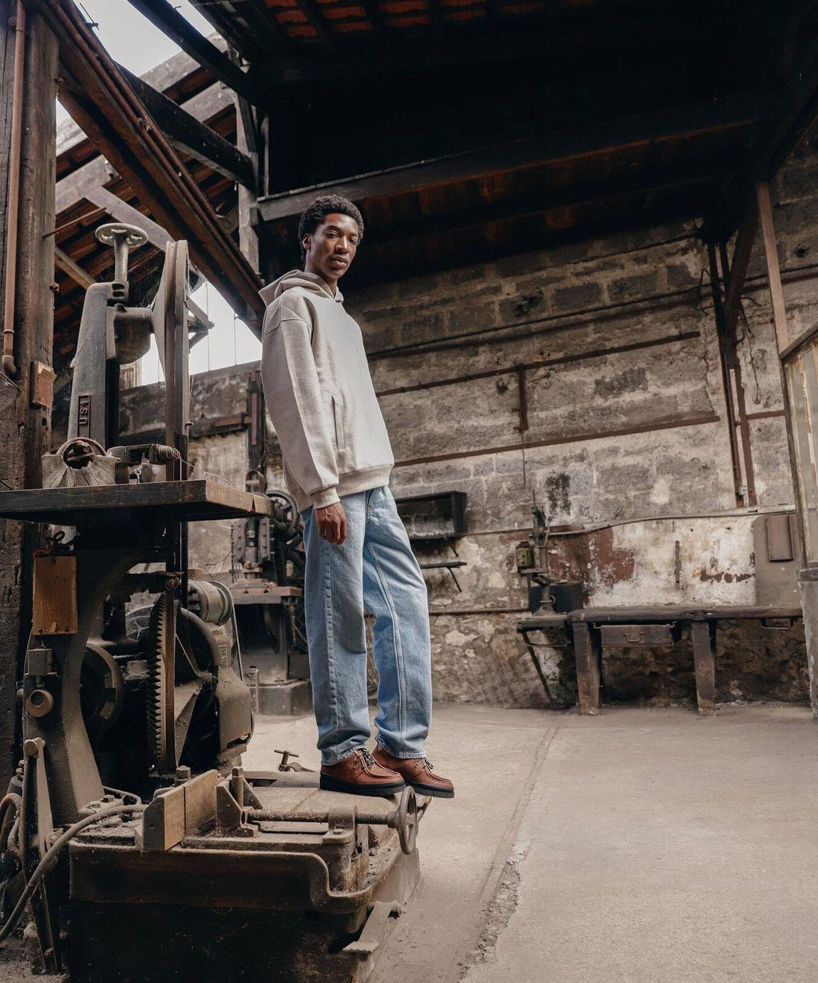 Kleman AW26 lookbook: a man in a grey hoodie and jeans standing in the Kleman workshop.