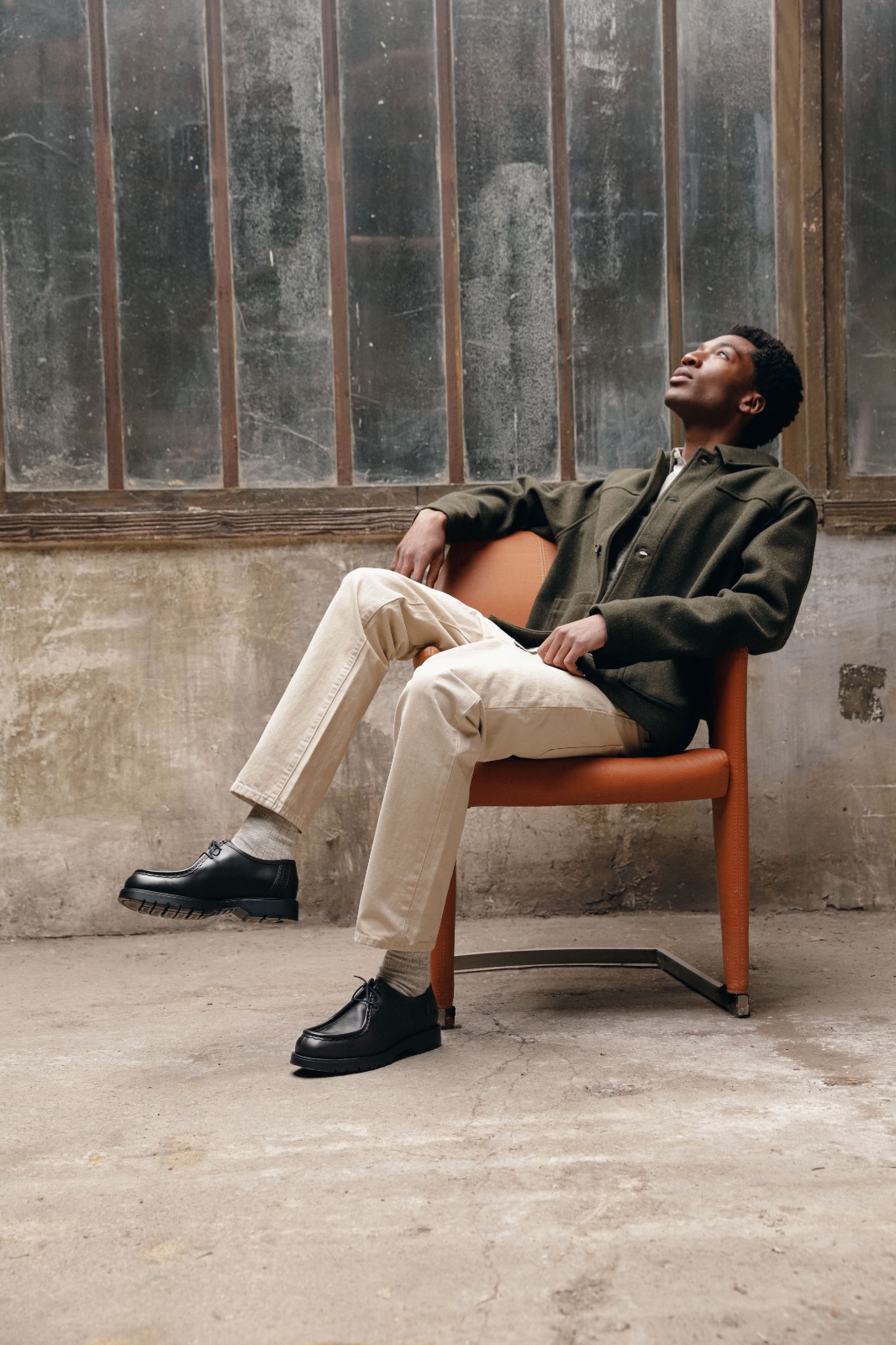 Kleman: a man in a dark green wool overshirt and cream trousers, seated in an orange leather chair, wearing black Kleman shoes, looking up at a weathered factory window.
