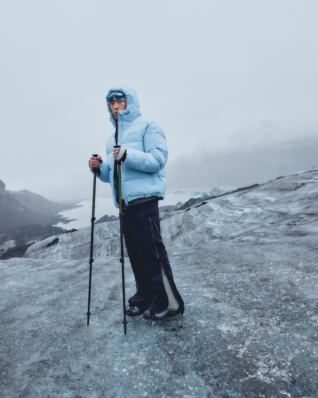 A woman in a pale blue down jacket and black trousers, with walking poles, on a glacier under low cloud.