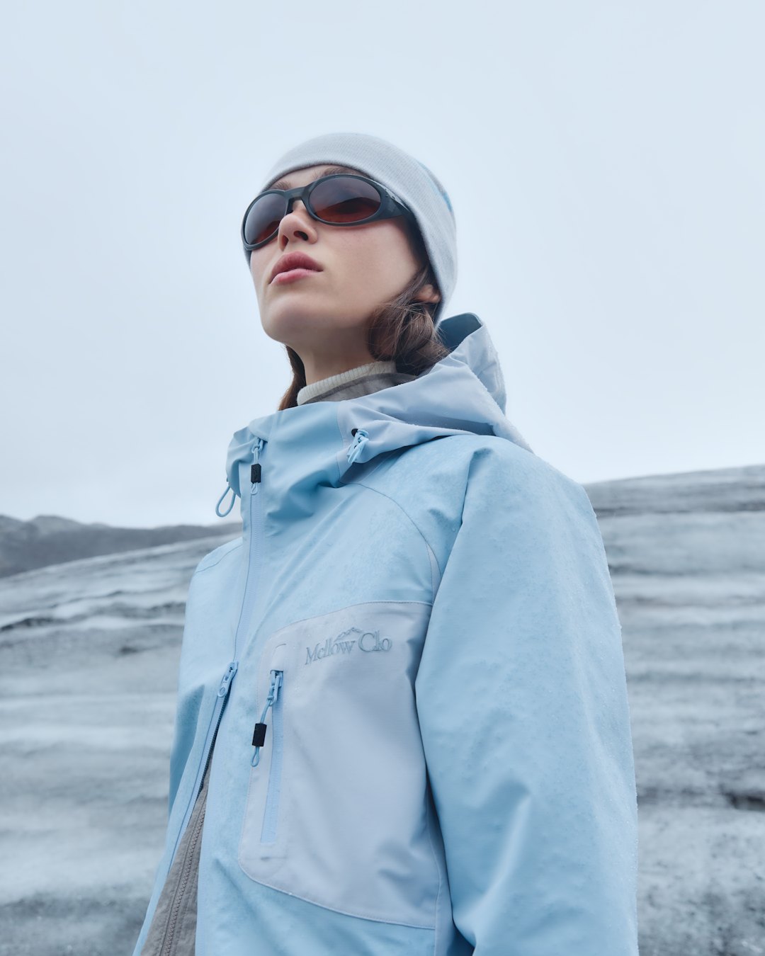 A woman in a pale blue hooded technical jacket, grey beanie and dark sunglasses, on a glacier.