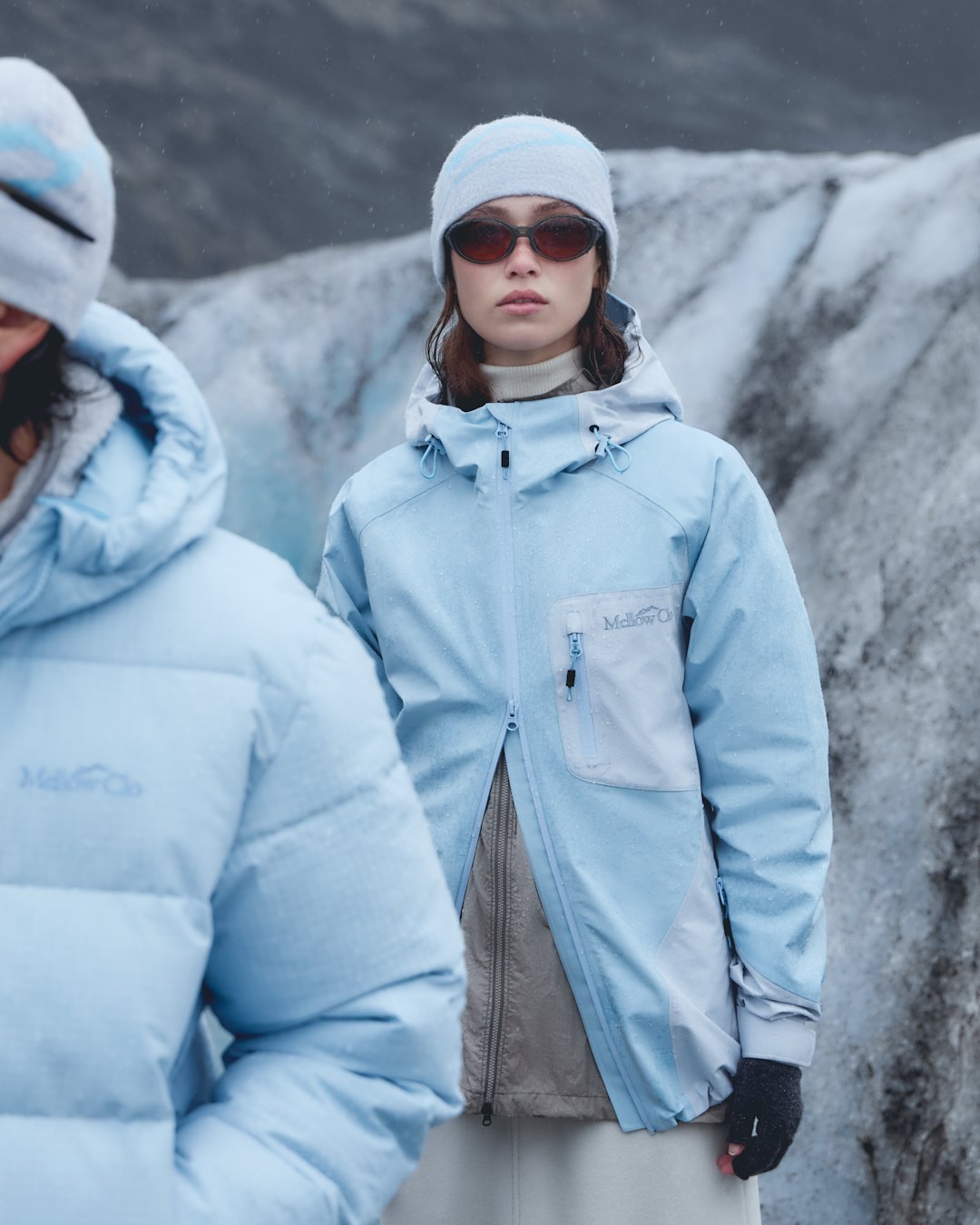 A woman in a pale blue hooded technical jacket with a grey chest panel, beside a figure in a pale blue down jacket.