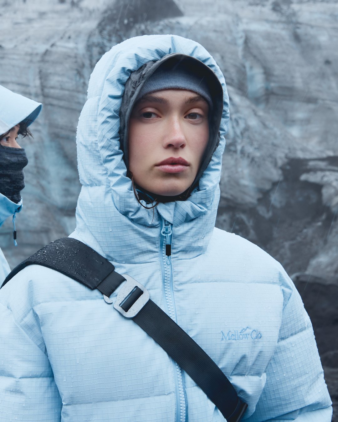 Mellow Clo: a woman in a pale blue hooded down jacket, the hood up, with a black bag strap across her chest, on a glacier.