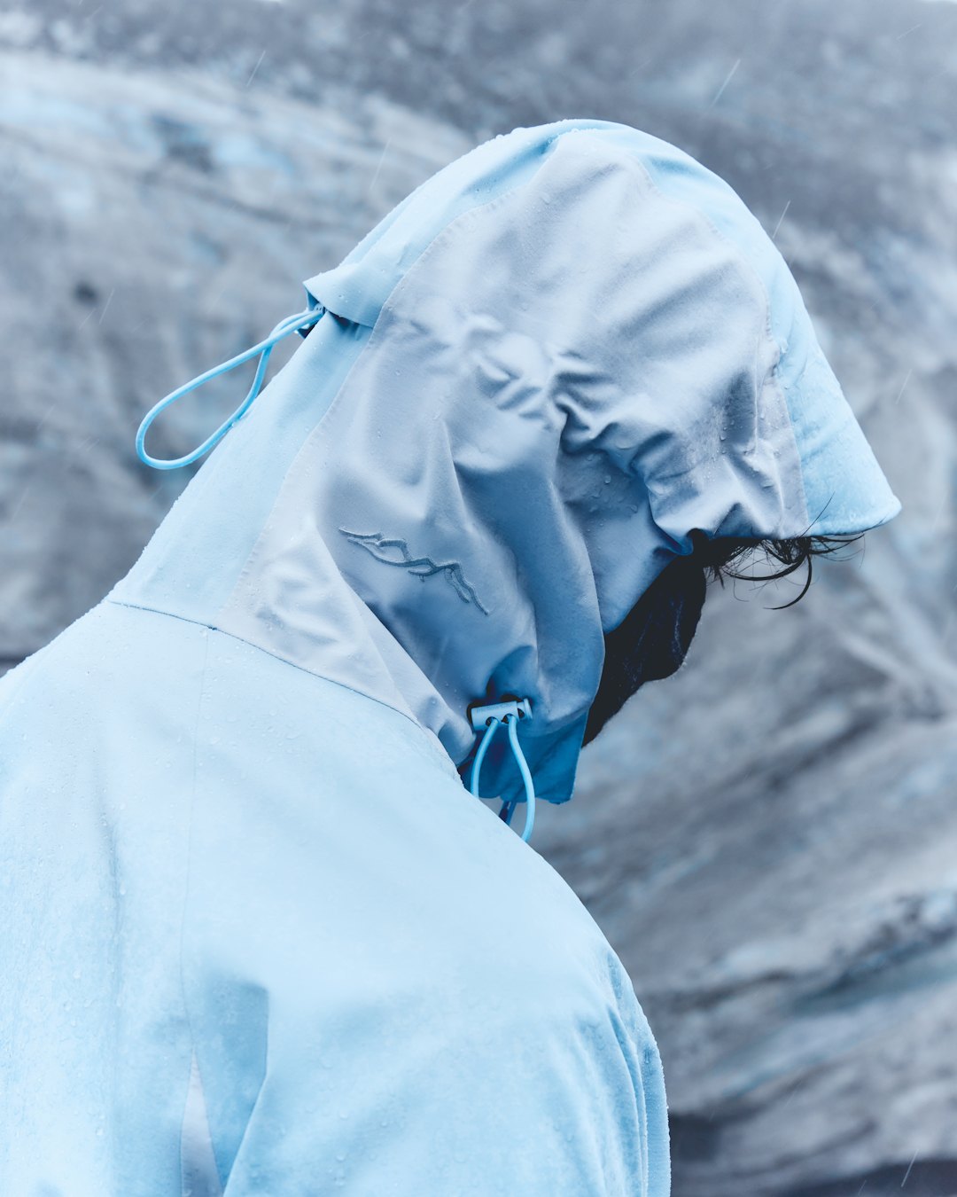 A pale blue technical jacket from behind, the hood up and its drawcord pulled.