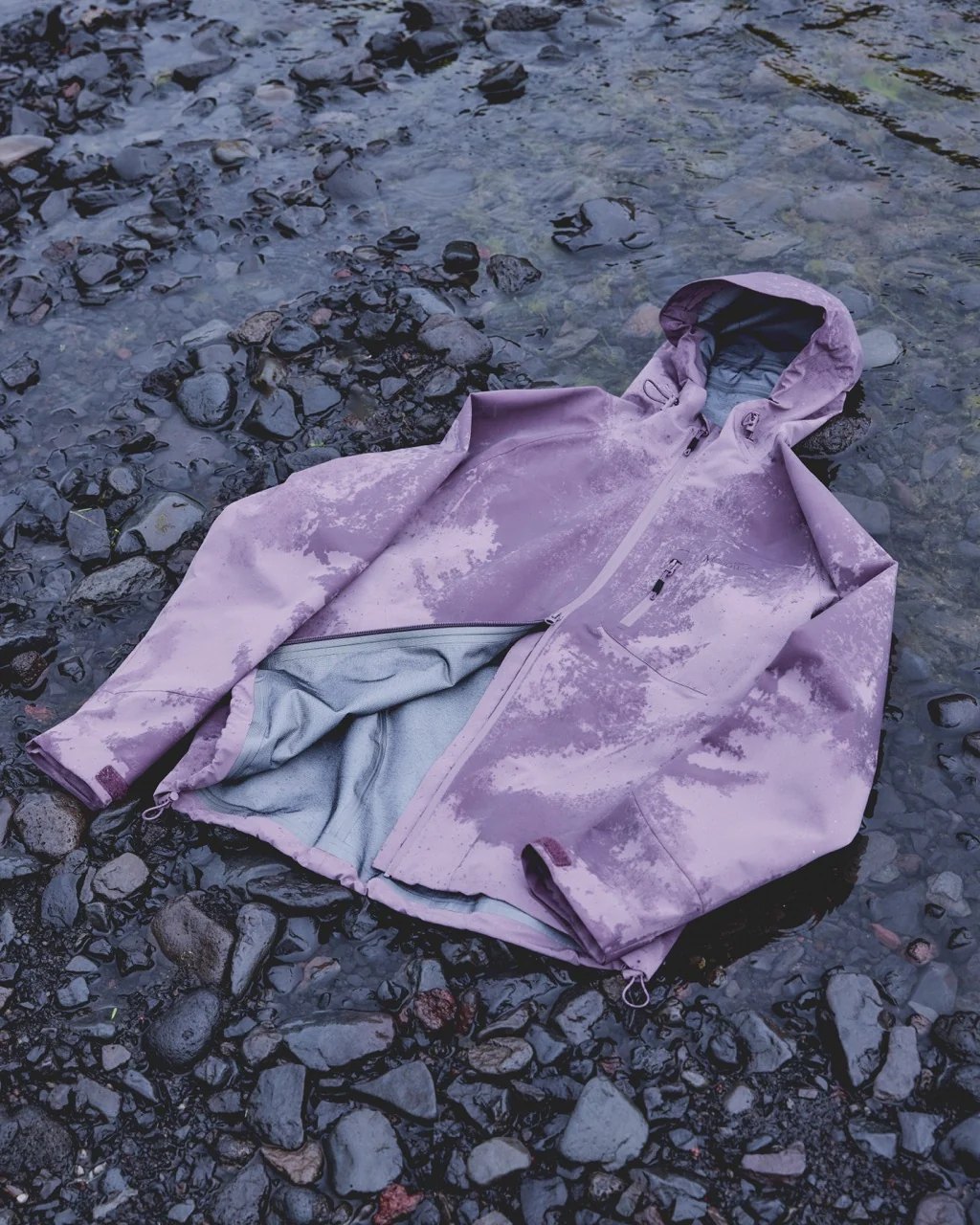 A lilac hooded shell jacket laid on wet stones.