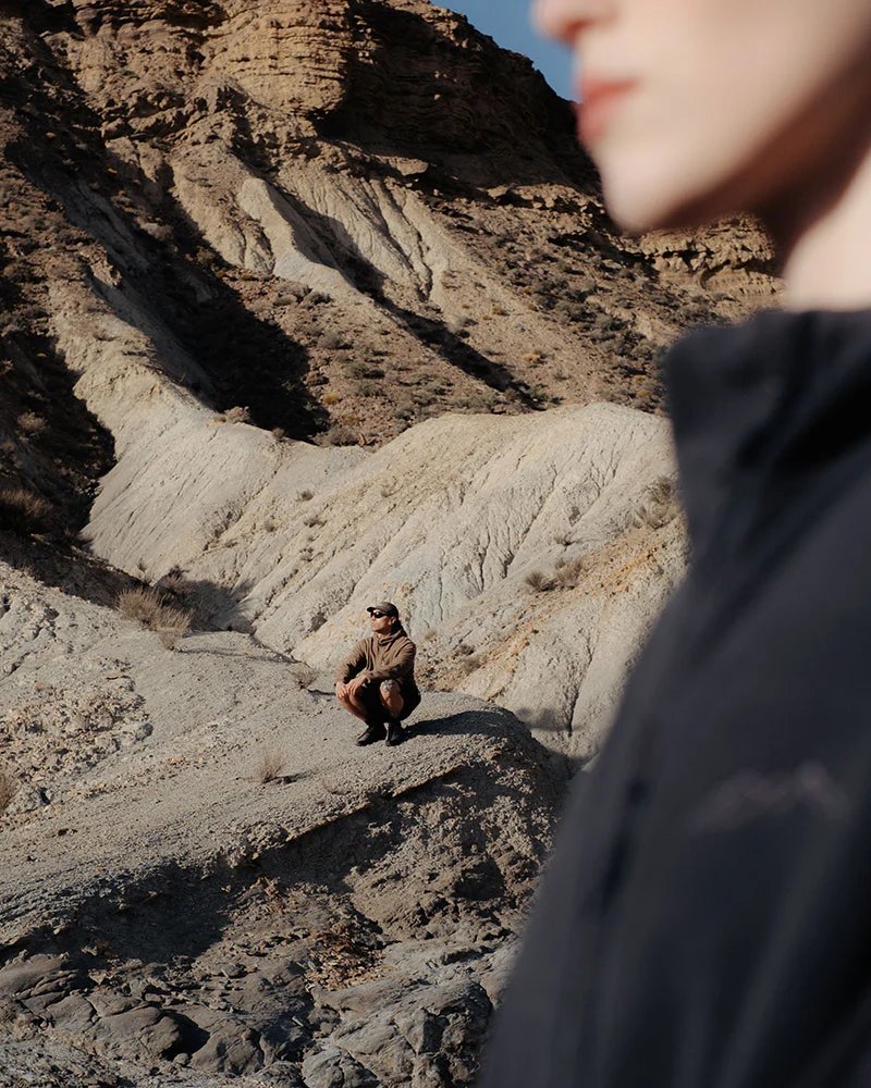 A man crouched in a dry canyon, with a figure in a dark jacket close to the camera.