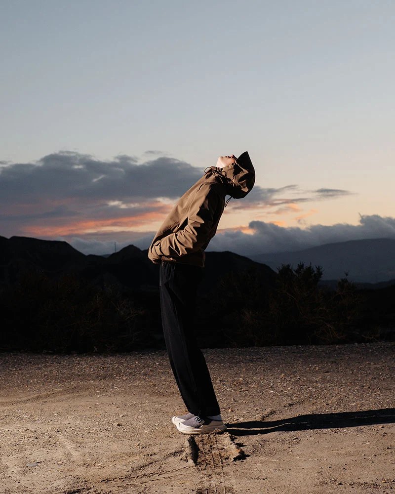 A man in a khaki jacket and black trousers stretching back against a dusk sky.