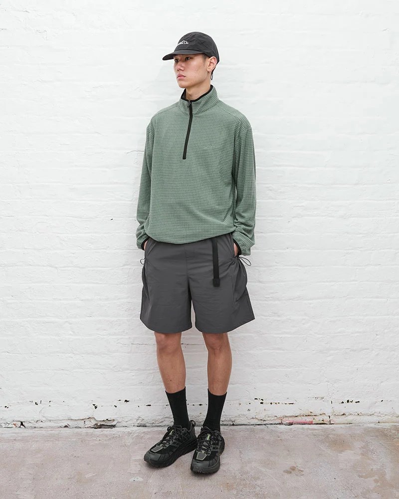 A man in a sage green half-zip fleece, grey shorts and black boots, against a white brick wall.
