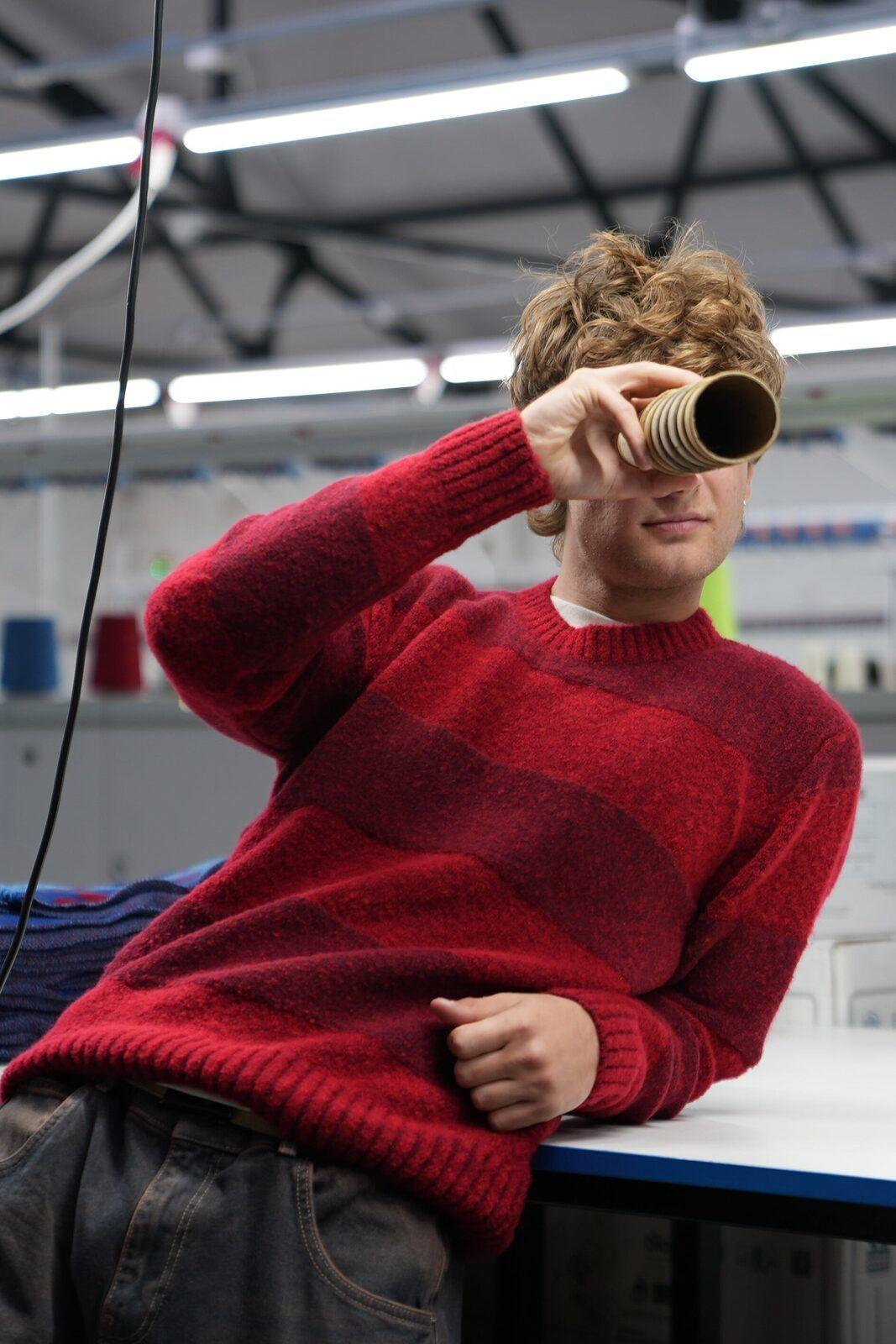 Origin: a man in a red striped jumper on the factory floor in Wigston, looking through a yarn cone.