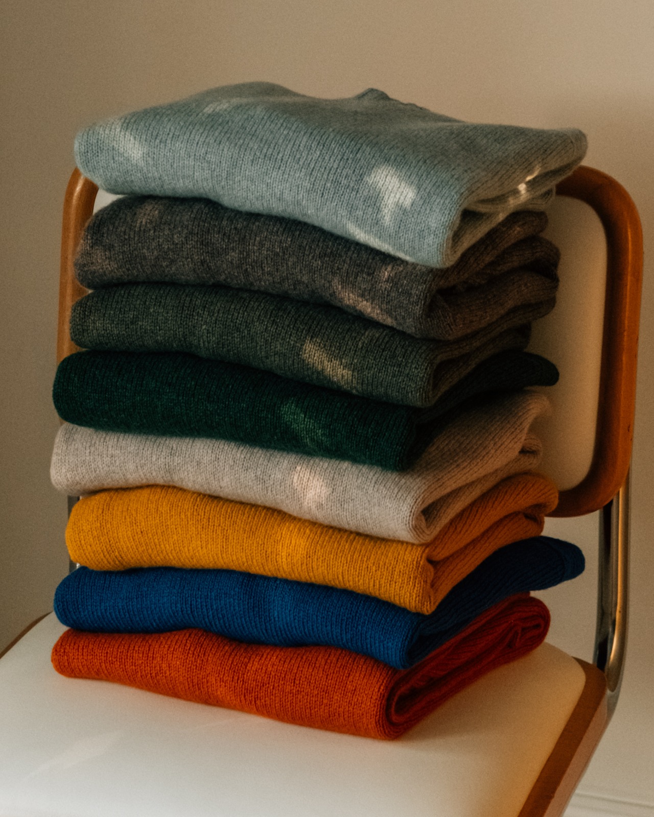 Origin: a stack of folded knitwear in pale blue, grey, green, cream, mustard, blue and orange on a chrome and leather chair.
