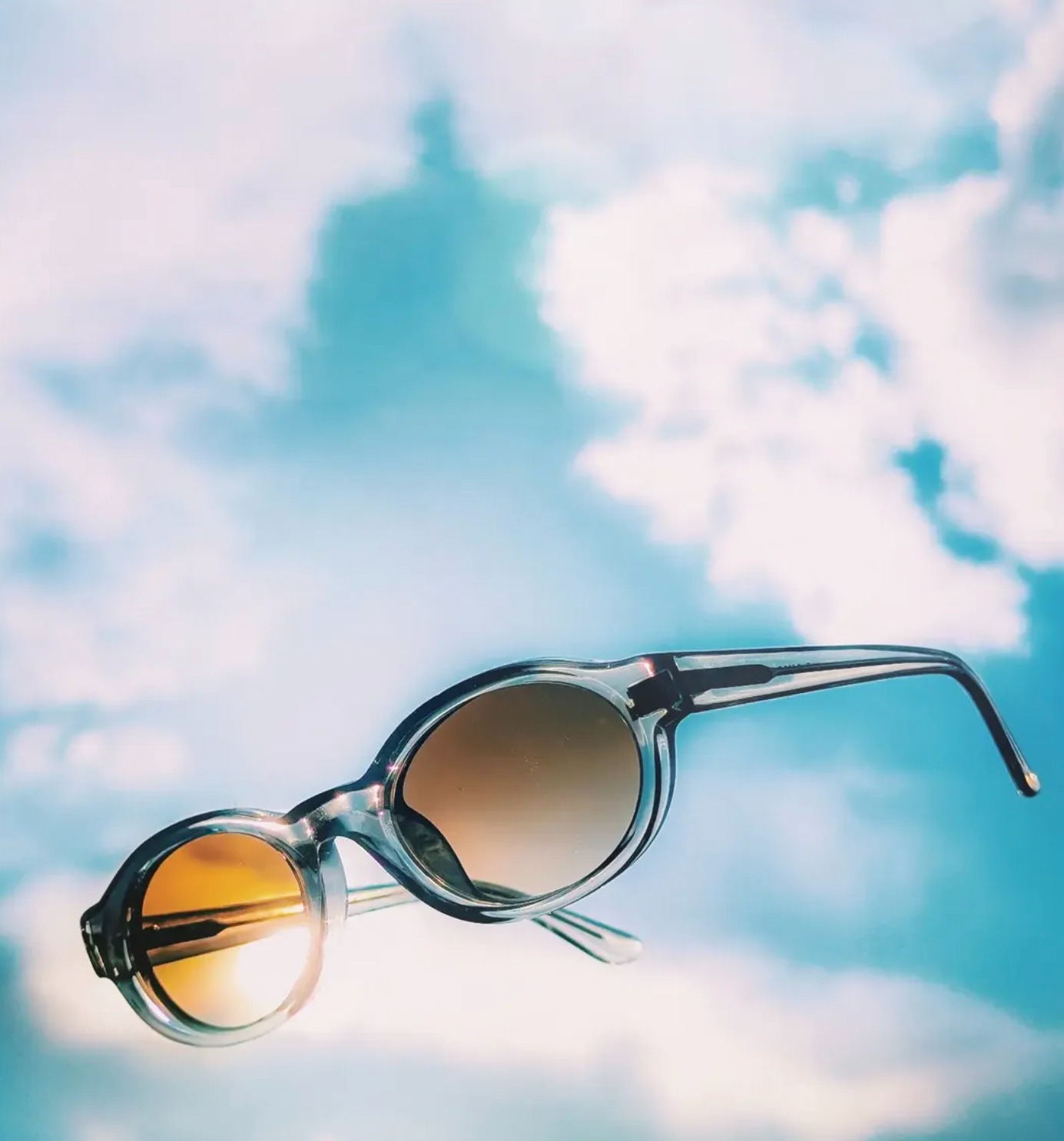 Waiting for the Sun: a pair of oval translucent-framed sunglasses with amber lenses floating against a blue sky and clouds.