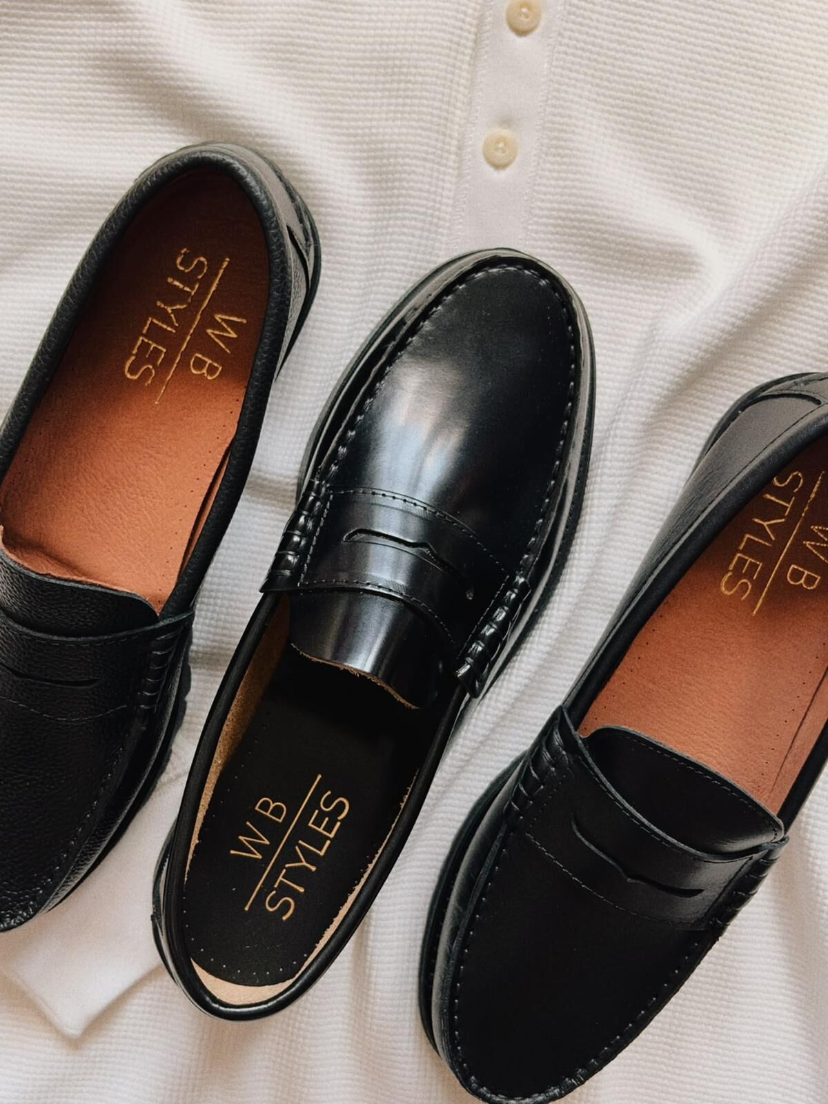 WB Styles penny loafers in black leather on a white knit, two showing the tan lining and the gold-stamped WB Styles insole.