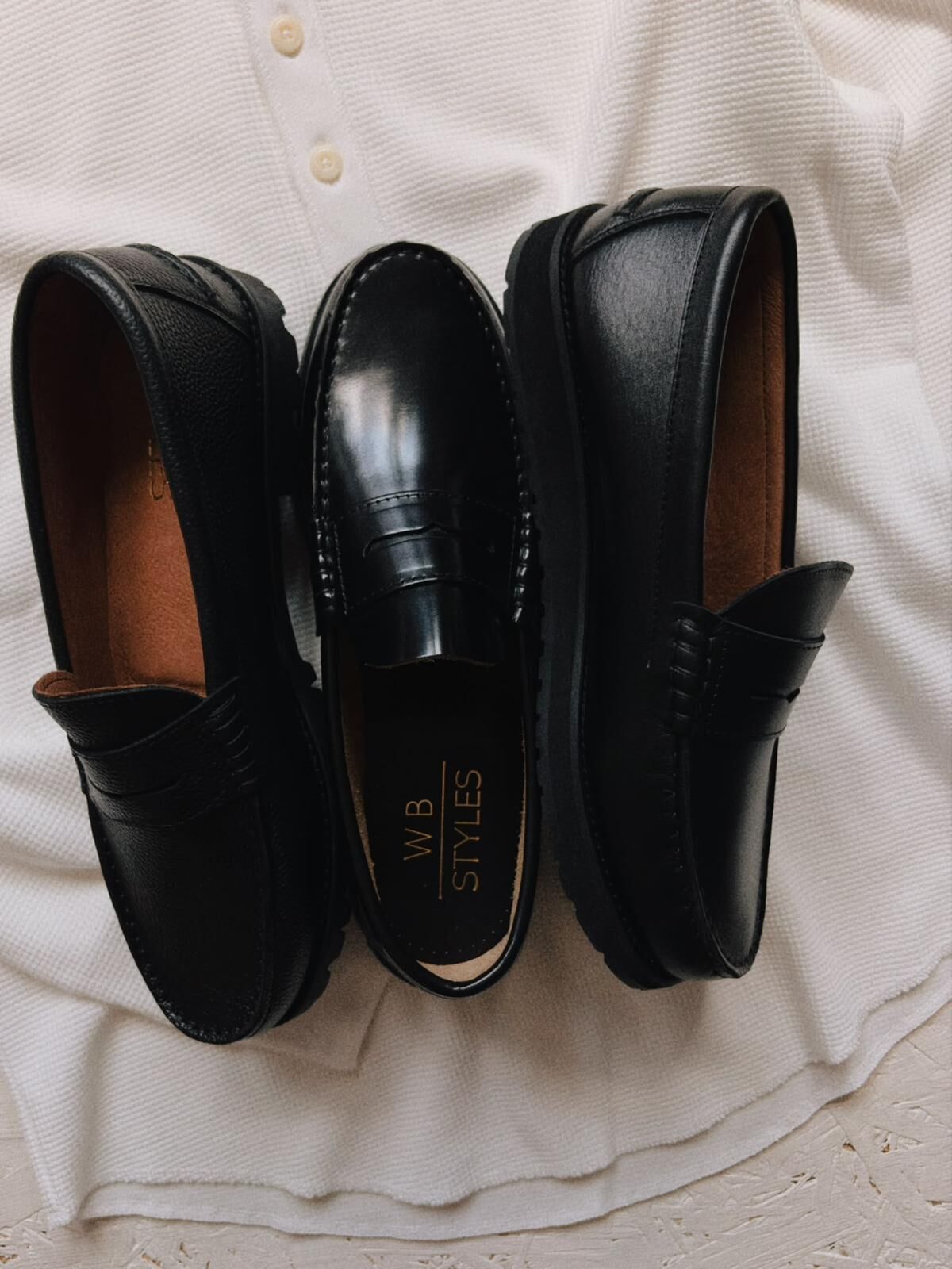 Three black WB Styles penny loafers side by side on a white knit, one showing the gold WB Styles insole.