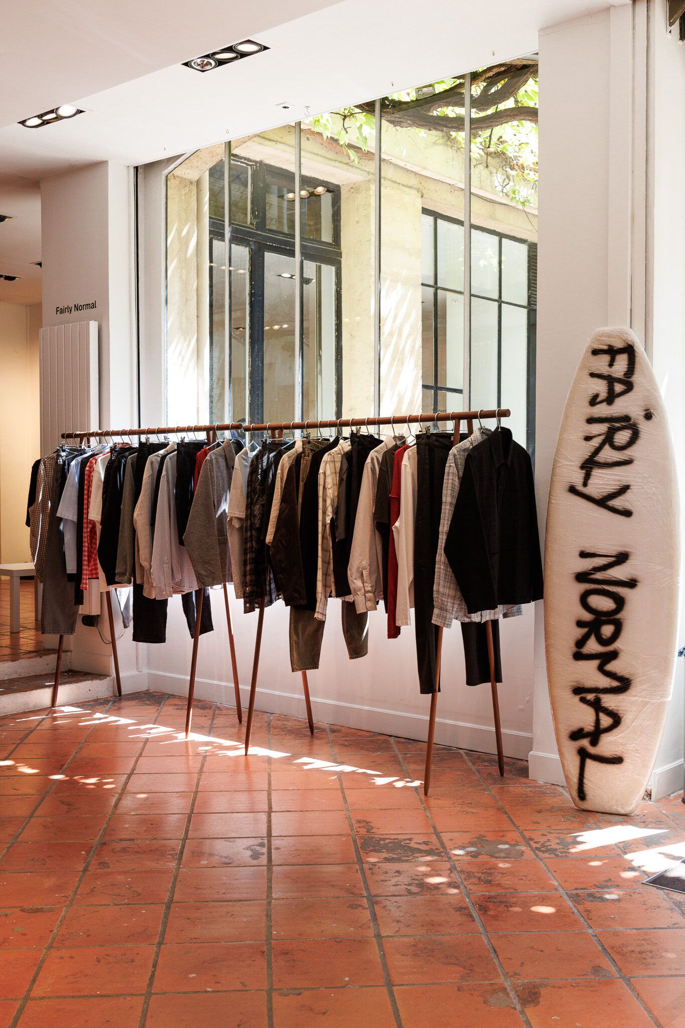 Rails of coats and trousers on the terracotta floor, a Fairly Normal surfboard against the wall.