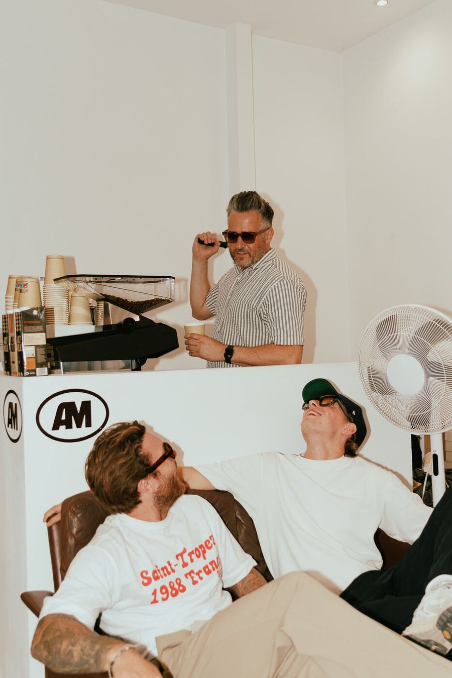 Two men laughing on a sofa beneath a fan, in the showroom.
